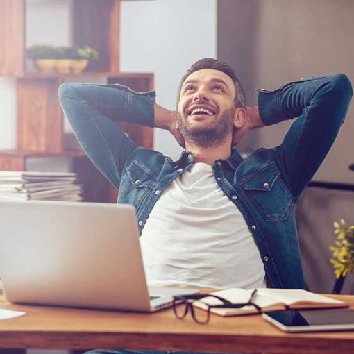Young man at computer, leaning back with hands behind head, smiling