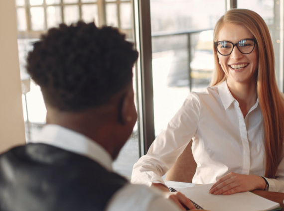 Coworkers sitting across from each other at a desk, smiling, writing tablets on desk. Photo by Gustavo Fring from Pexels.com.