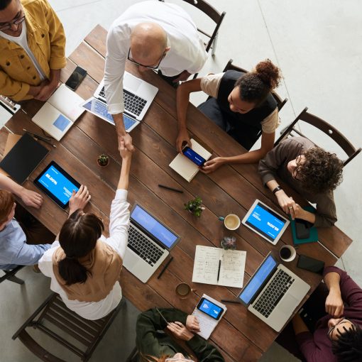 Photo of eight people at an office table with computers, phones, and tablets. Two people, man and woman, shaking hands across table.