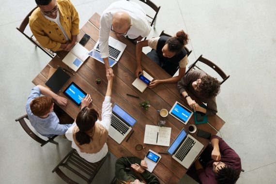 Photo of eight people at an office table with computers, phones, and tablets. Two people, man and woman, shaking hands across table.